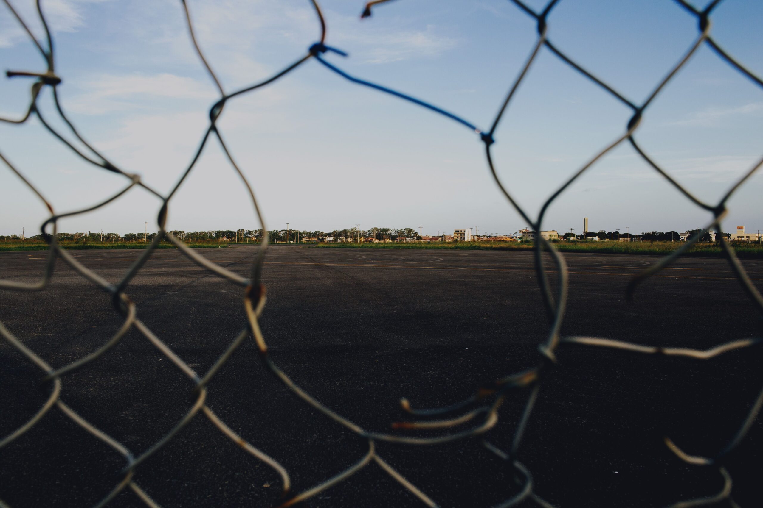 A broken wire mesh fence framing an open asphalt area under a blue sky.